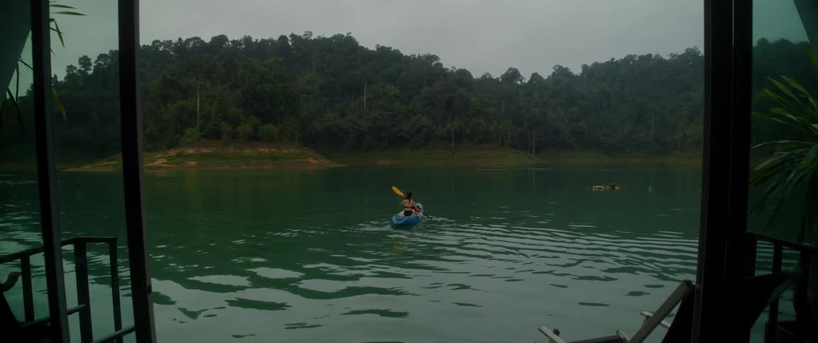 A woman slowly paddles a kayak away from a riverside house