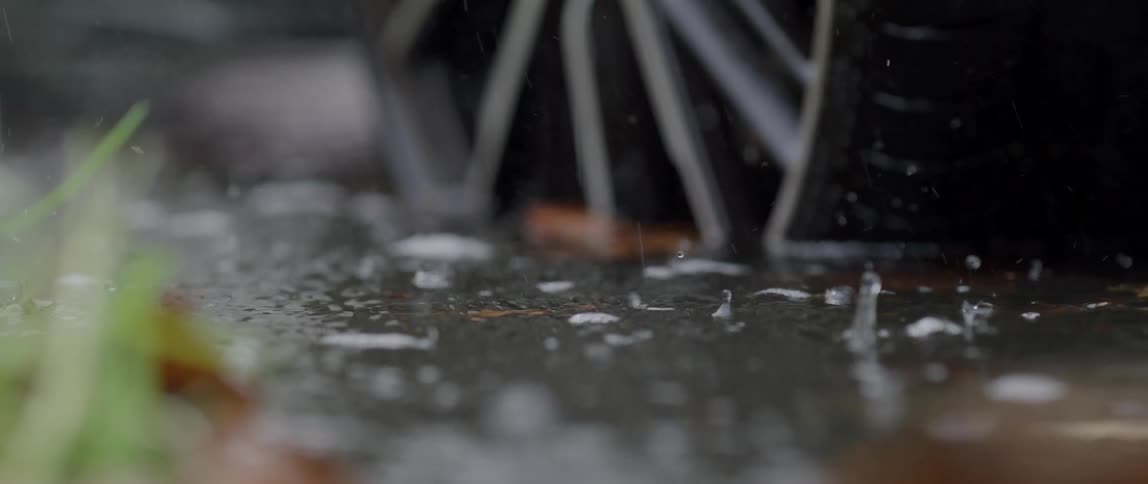 Slow motion closeup of raindrops falling on the ground and creating splashes of water with a vehicle wheel in the background