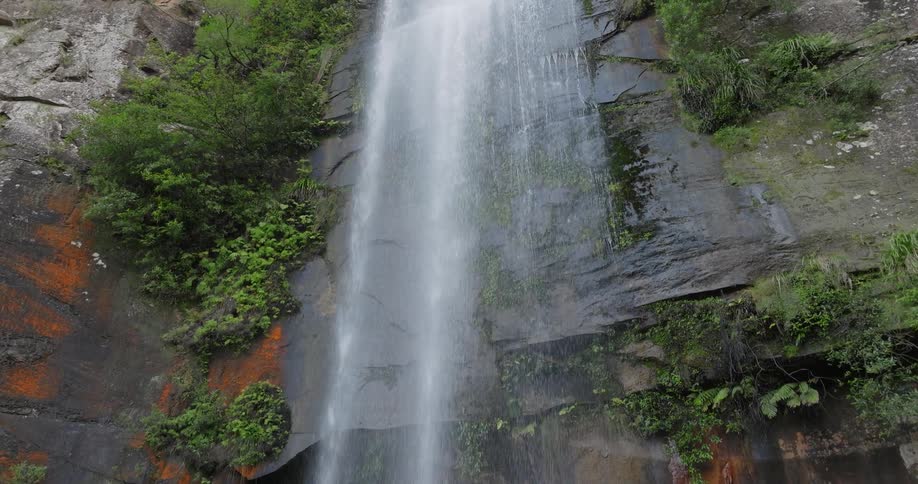 Plants and vegetation grow along the side of a steep cliff as waterfalls flow into the river below