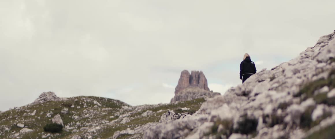 Blonde woman walking on a trail with her back to the camera beneath the mountains on a cloudy day