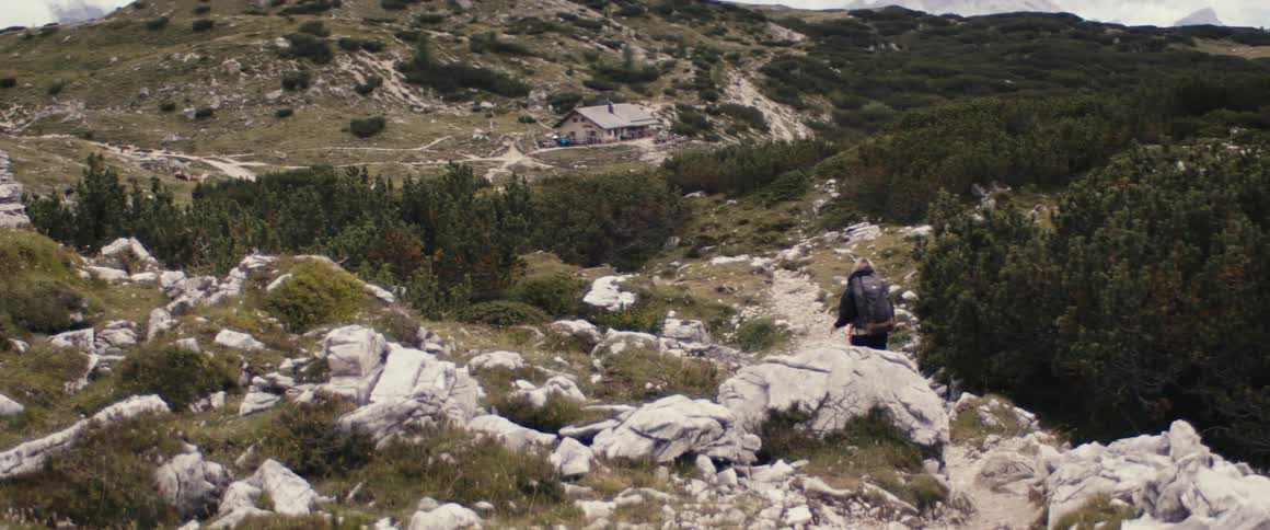 Backpacker hikes along snowy trail up slope toward lone house