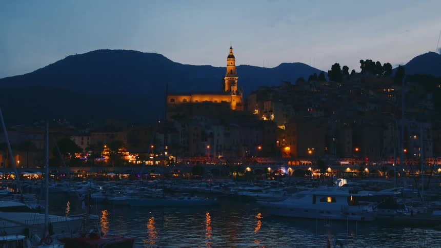 Docked yachts and in the background an illuminated church at night with a mountain range on the horizon