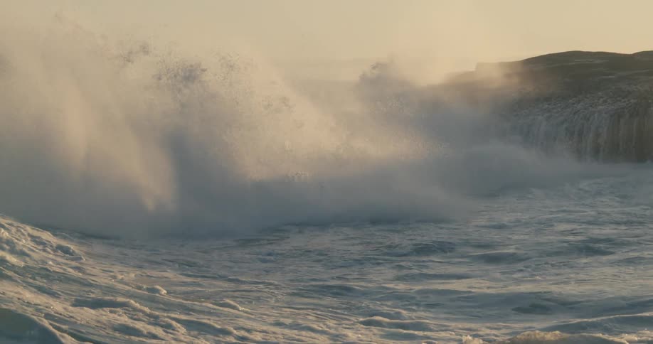 Drone view of waves crashing over the ocean surface onto rocks on a bright sunny day