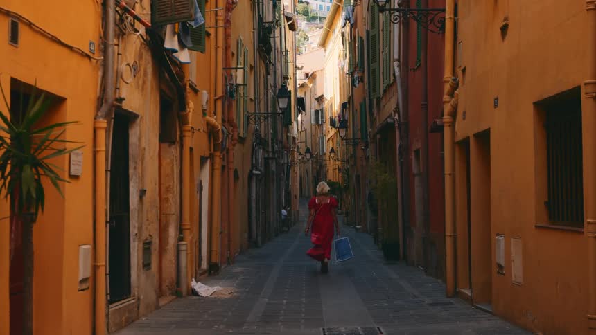 A woman wearing a red dress carries a shopping bag as she walks on an alley surrounded by tall orange buildings