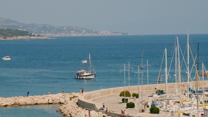 Docked boats and others in motion on the sea with people walking on a sunny day