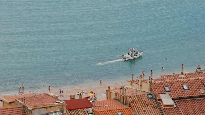 Boat moves away from busy beach near red roofed buildings