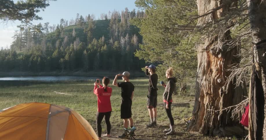 A group of men and women stand outside a campsite by the water taking pictures and observing