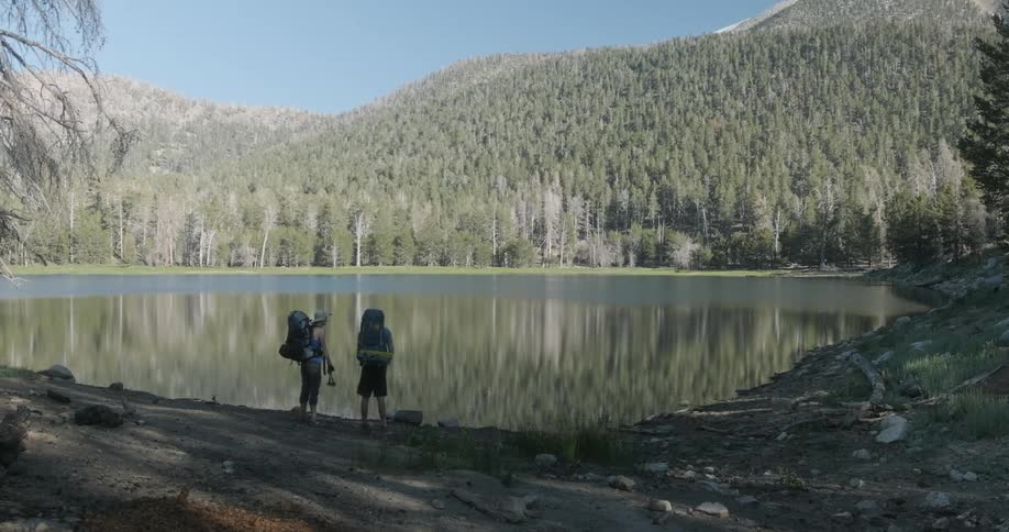Two people wearing large packs stand at the edge of a lake surrounded by hills