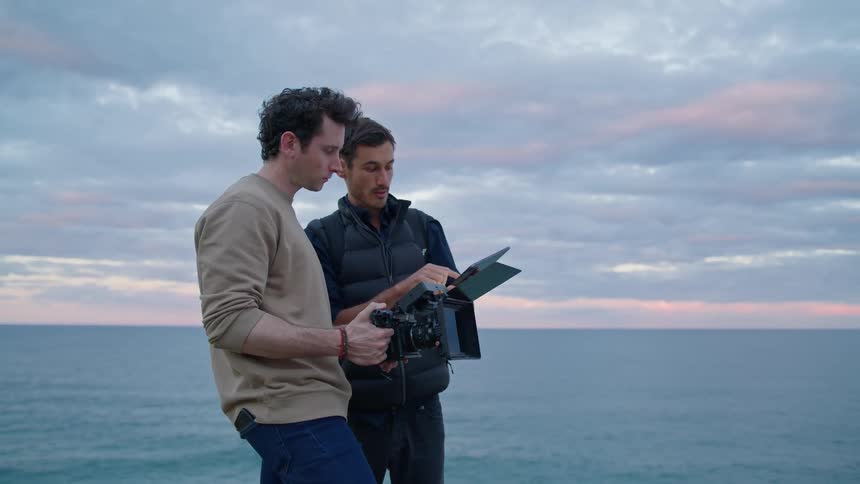 A man holding a camera and a man with a table computer are standing along the coast looking at their equipment and talking