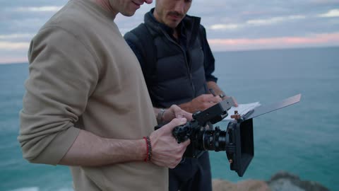 Man setting up a professional camera next to another man who is observing and holding a pen both atop a seaside coast