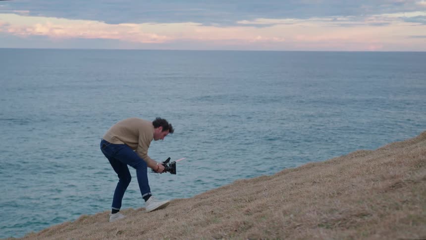Photographer stands downhill of man on coastal hill