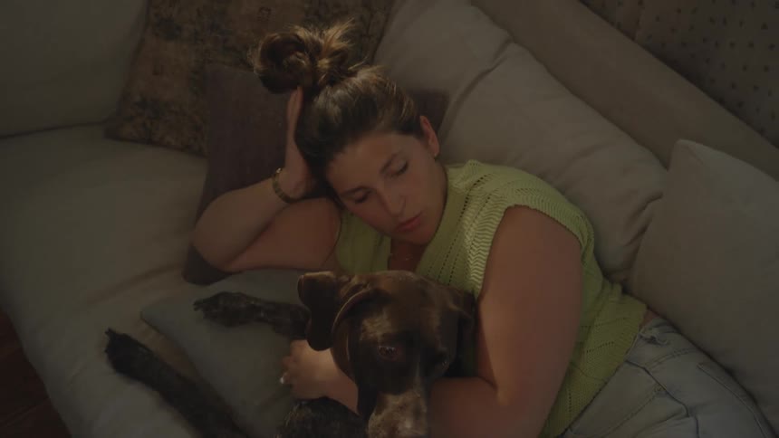 Young woman indoors laying on a couch in a living room petting a dog