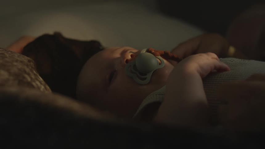 A baby lying in bed sucking on a pacifier as a parents hand caresses their head