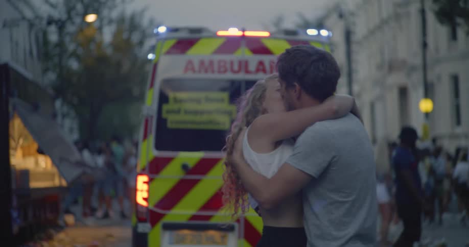 Man and woman embrace in front of ambulance as it pulls away