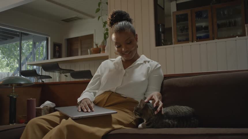 Woman seated on the sofa in formal attire holding a laptop with a cat being caressed by her side