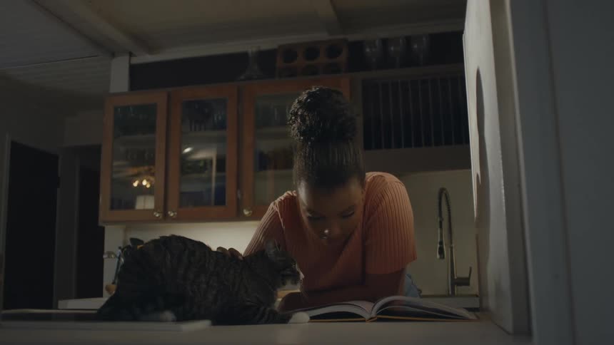 A teenage girl is standing in the kitchen looking at an open book on the counter as she pets her cat