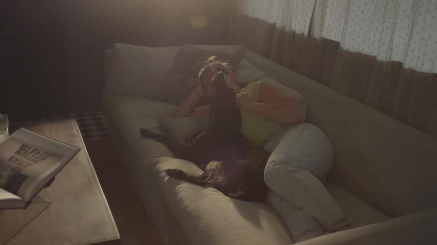 Woman lying down on a couch indoors petting a brown dog during day time