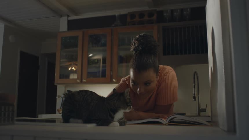 A teenage girl is standing in the kitchen with an open book on the counter as she pets her cat