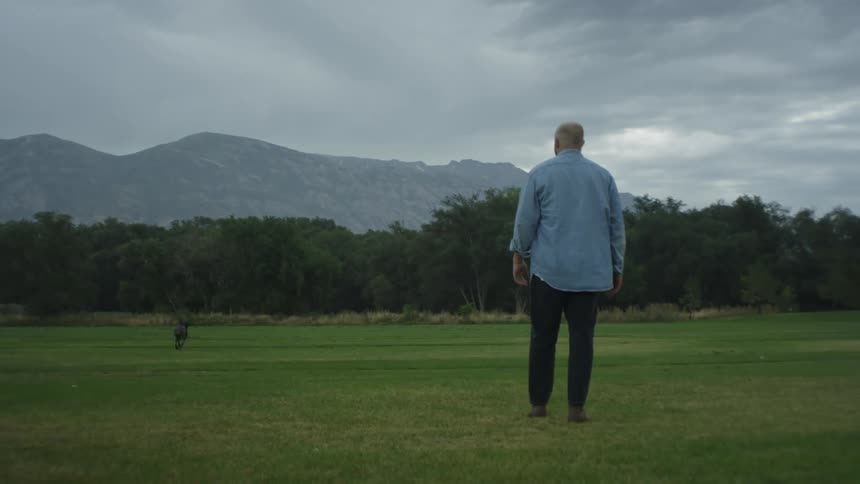 Man throwing a stone as his dog runs to fetch it in an outdoor setting with grass trees and a chain of mountains on the horizon on a cloudy day