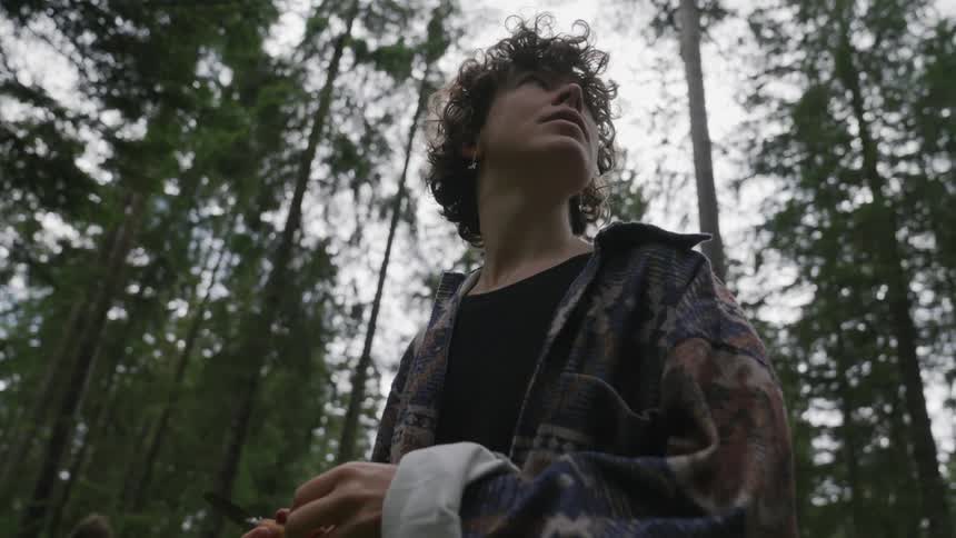 A woman in coats in the middle of a forest of large trees looking around