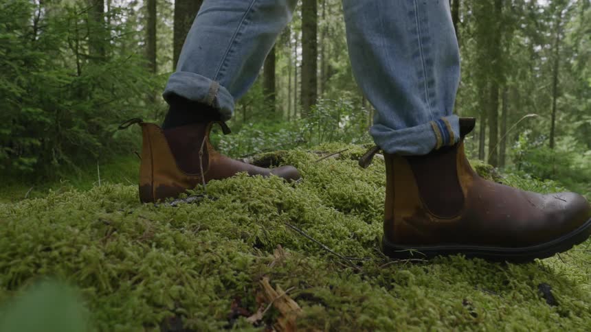Legs with cuffed jeans wearing brown leather boots walking on a mossy slope in the woods