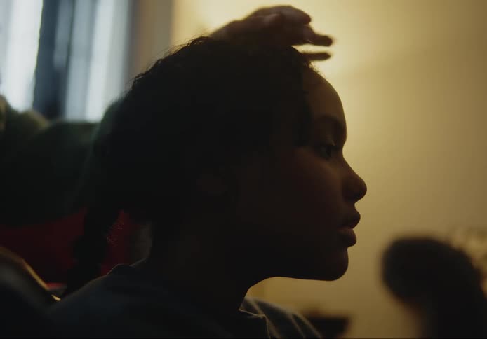 Close up of a young girls face while she has her hair braided indoors in a room lit with a lamp