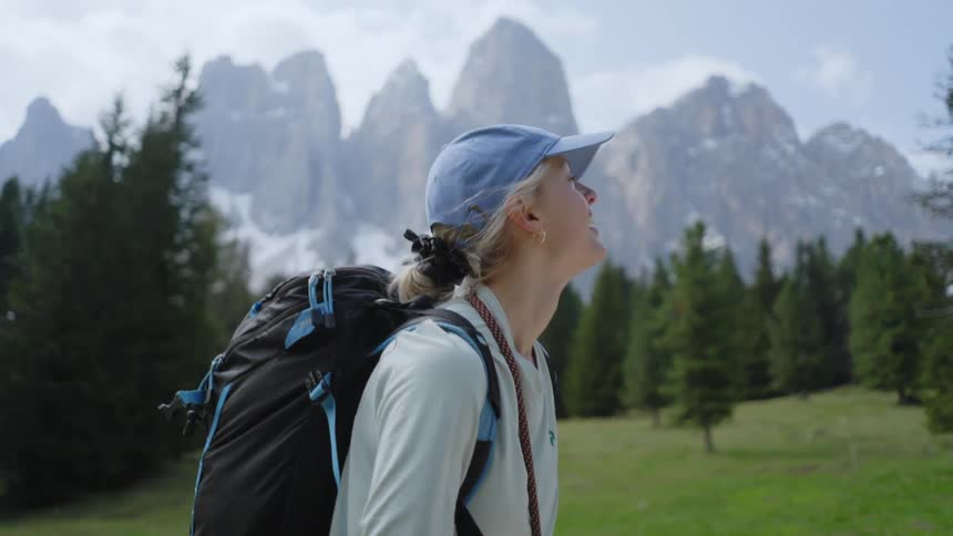 A woman hiking through nature in a forest of pine trees with tall mountain peaks behind her