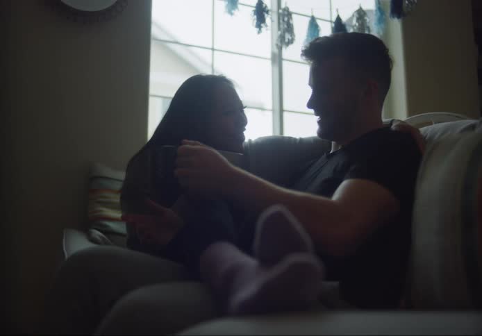 A man and a woman sit on a sofa with mugs in their hands while smiling and talking