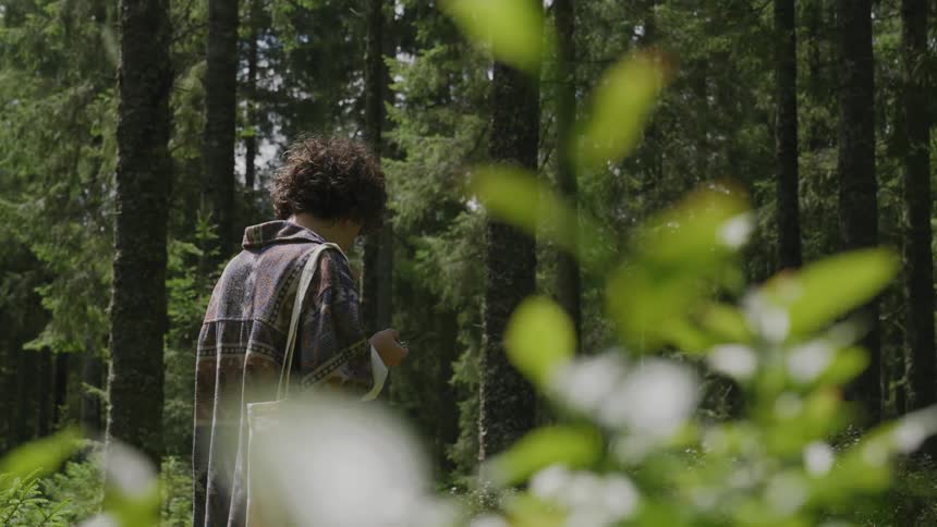 A person in a sweater eating something walking in the middle of the forest with a bag around his waist