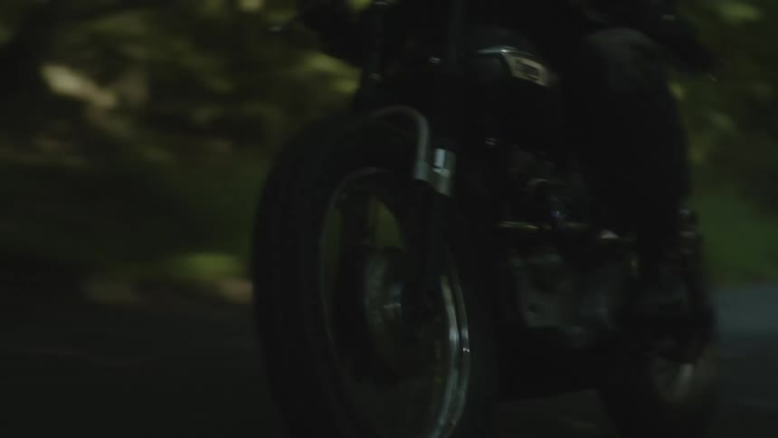 A biker in a leather jacket and black helmet riding a motorcycle near the trees