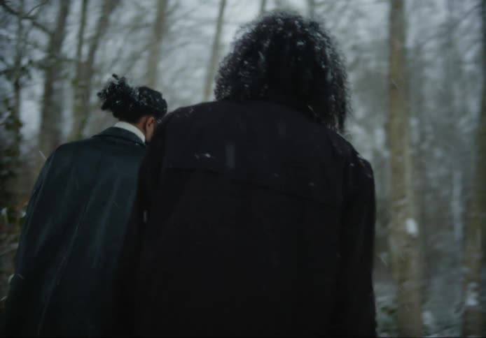 Two men wearing masks walking in the woods in the snow during the day while holding a camera