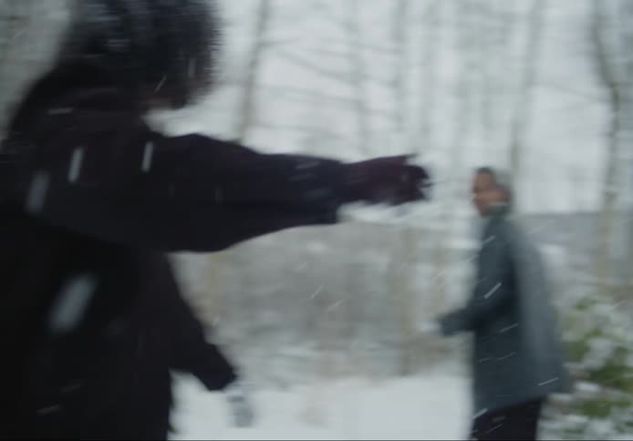 Two black friends playing outdoors by throwing snowballs at each other next to a forest with houses in the background during day time
