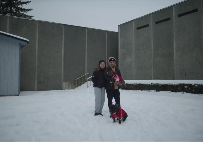 A man and a woman are outside standing together in the snow as he holds a dog in his arms and another dog is sitting in front of them