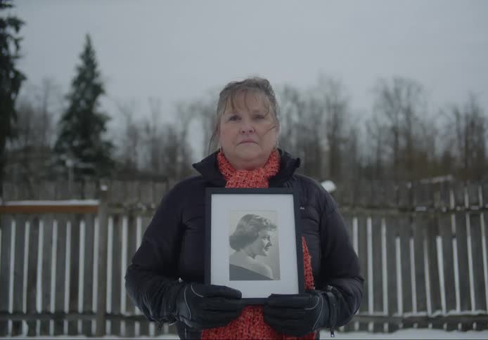 Elderly woman outdoors on the snow standing and holding a black and white picture of another woman in her hands next to a fence and a forest during day time