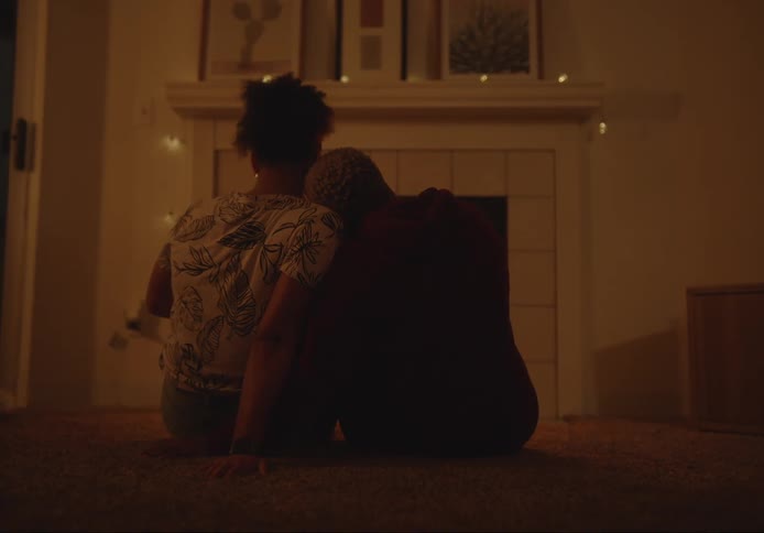 Two women sitting on the floor back to back with one resting her head affectionately on the shoulder of the other