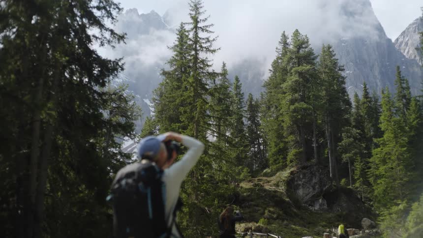 A woman wearing a backpack taking a picture of tall pine trees at the base of tall mountain ranges with fog settled in front of them