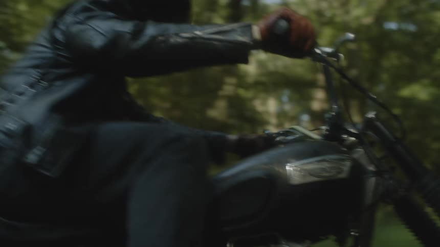 A biker in a leather jacket and black helmet riding a motorcycle near the trees