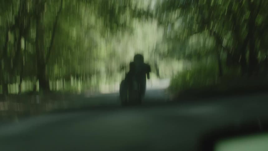 A man is driving an automobile on a road through the woods as a motorcycle passes his car and speeds in front of him