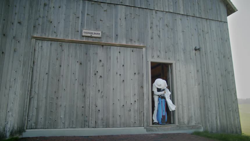 A man in a hat with all white clothes coming out of a barn and touching a white cloth