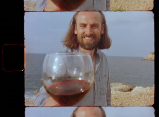 A man smiles and expresses happiness and gratitude as he clinks wine glasses in a toast with his friend or partner standing on a hill overlooking the ocean filmed on Super 8 mm in a POV or FPV perspective