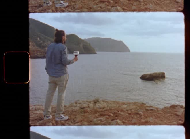 Man in gray shirt and beige trousers holding a red wine glass and enjoying the ocean view as the sun is setting at the horizon over the sea while he enjoys a silent and peaceful moment on his own on Super-8 film with retro and nostalgic feeling