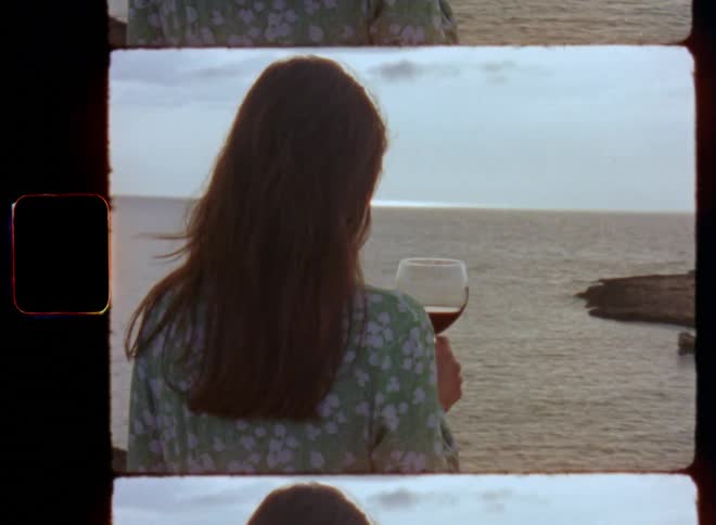 A lonely or lonesome brunette woman wearing a green flower dress with a glass of wine watches the sea at sunset while standing alone on a cliff contemplating in sad moment as she reflects her life while enjoying the view of the setting sun at the horizon