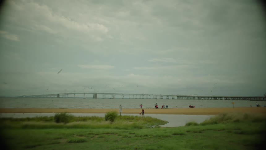 Landscape of people on the sandy shore of a sea with a bridge in the background