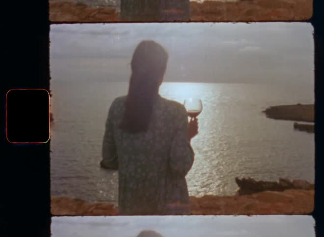A brunet woman in a green flowered dress swaying in the wind or breeze on the shore of the beach at the sea holds a glass of red wine as her boyfriend approaches her and places his hand on her shoulder touching her to express fondness and love