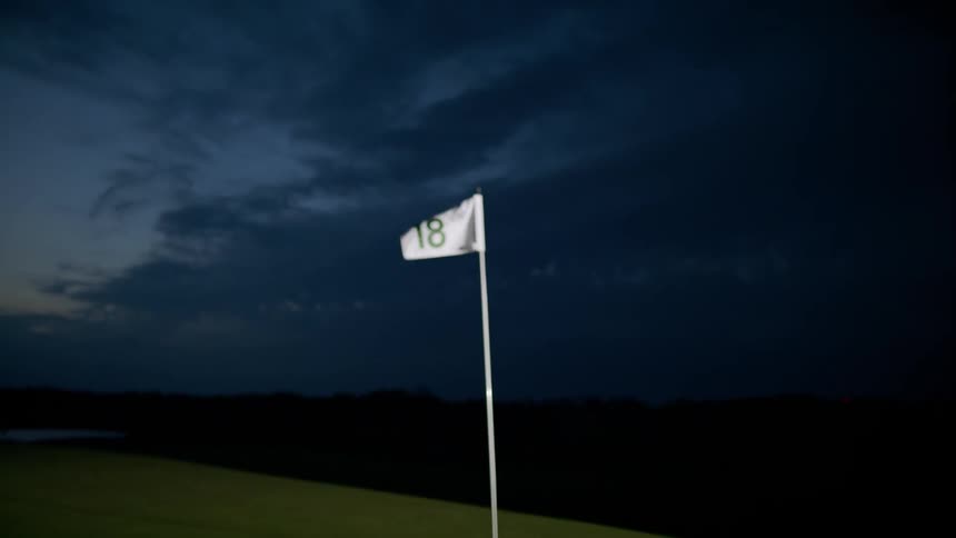 A flag with the number 18 stamped on it is planted on a golf course and is moving with the wind