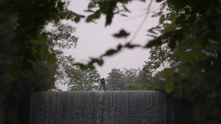 Two people are walking toward each other across the top of a waterfall and they meet in the middle and hug