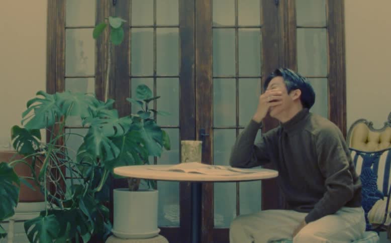 A man is sitting at a table near potted plants as he opens a magazine and laughs putting his hand on his face