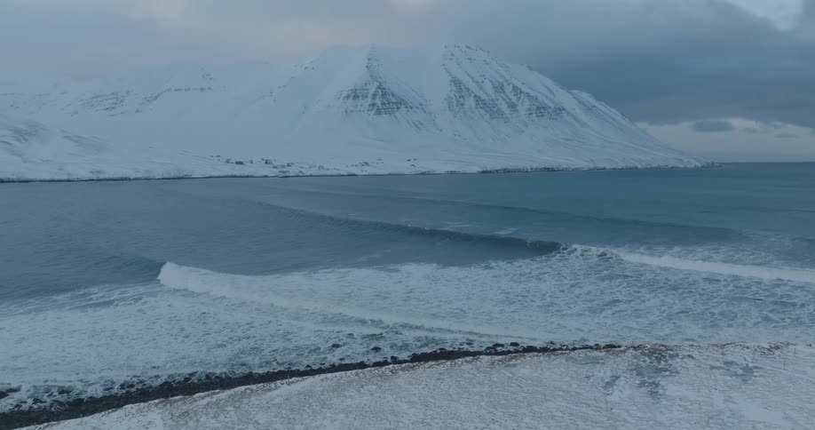 Aerial of ocean waves near the coast next to snow covered mountains