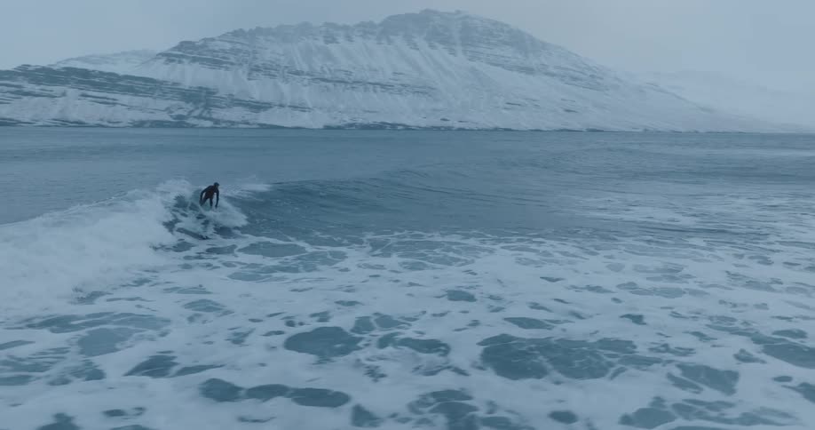 A surfer in a wet suit rides a wave in an area with snow covered mountains