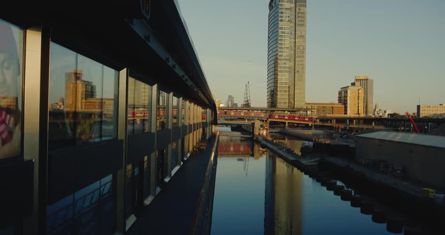 A picture of a man and nearby buildings reflect on the windows of a building along a waterway as a train moves over a bridge by a tower and buildings in the distance